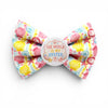 A novelty dog bow tie featuring a colourful seashell and starfish print on a striped background, with a round white centre badge that reads 'THE WORLD IS MY OYSTER'.