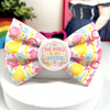 A pastel seashell patterned dog bow tie with a central round white badge that reads 'THE WORLD IS MY OYSTER.' The bow tie is displayed on a white stand against a softly blurred indoor background with colorful decor.