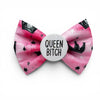 A pink bow tie for dogs with a watercolour texture, featuring black crowns, stars, and dots, with a white circular badge in the centre that reads 'QUEEN BITCH' in black text.