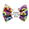 A Halloween inspired dog bow tie featuring a vibrant, multicoloured print of cartoonish monster eyes, with a white circular centre patch that reads 'KEEPING MY EYE YOU BITCH' in black text.