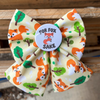 A woodland fox print dog bow tie, on a pale yellow background, featuring green trees and orange foxes, secured with a round white button that reads 'FOR FOX SAKE' with a small fox illustration.
