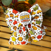 A white fabric dog bow decorated with a colourful autumn pattern of acorns, mushrooms, pumpkins, leaves, and coffee cups, featuring a round badge in the centre that reads 'I'M SO CUTE EVEN THE LEAVES FALL FOR ME'. The bow is displayed on a wooden surface with decorative plants and a glass bowl in the background.