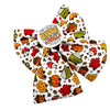 A autumnal dog bow tie featuring a white background with a colourful autumn-themed cartoon print of pumpkins, acorns, mushrooms, leaves, and a coffee cup. A circular badge tag is attached to the centre with the text 'I'M SO CUTE I SEND THE LEAVES & FALLS TO ME'.