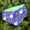 A sweary Halloween dog bandana. Adjustable, with a purple Halloween print featuring ghosts, tombstones, and the text 'Spooky Little Fucking' is shown worn around a green leafy plant. The inside of the collar is light green with white snap buttons.